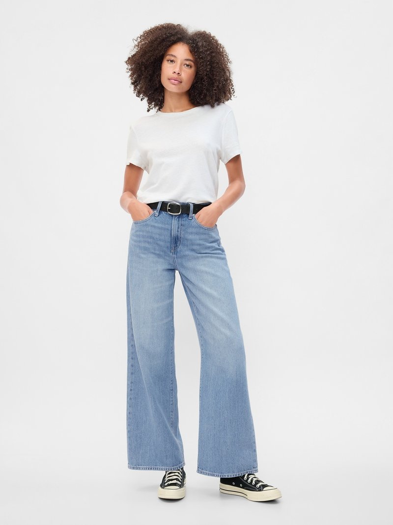 Junge Frau mit lockigem Haar, die ein weißes T-Shirt, weit geschnittene blaue Jeans mit schwarzem Gürtel und schwarze Sneaker trägt, steht mit den Händen in den Taschen.