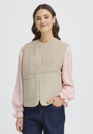 Kobieta nosząca beżową kamizelkę polarową na zamek na różową bluzkę z długim rękawem oraz niebieskie dżinsy, stoi z jedną ręką w kieszeni, uśmiechając się.