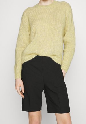 Osoba v světle žlutém pleteném svetru s dlouhými rukávy a černých krátkých šortkách na kolena, s jednou rukou v kapse.
