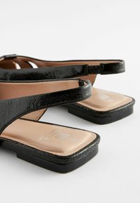 Schwarze Ledersandalen mit strukturierter Oberfläche, einem niedrigen quadratischen Absatz und breiten Riemen. Die Fußbetten sind beige und glatt, mit eingeprägten Markeninformationen.