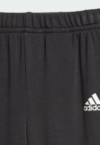 Pantaloni della tuta neri in morbido tessuto con cintura elastica, dotati di logo Adidas bianco sul lato sinistro. Materiale elastico, texture liscia.