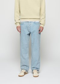 Person iført lyseblå rette jeans, beige sweatshirt, hvit skjorte i kanten, og beige joggesko med brune striper, stående foran en hvit bakgrunn.