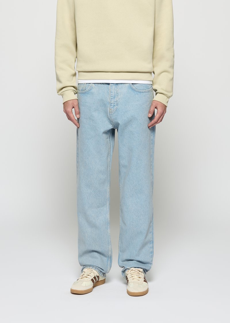 Person iført lyseblå rette jeans, beige sweatshirt, hvit skjorte i kanten, og beige joggesko med brune striper, stående foran en hvit bakgrunn.