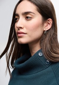 Teal cowl neck tröja i texturerat tyg, med två mörka knappar på kragen och bärs med silverfärgade hoopörhängen.