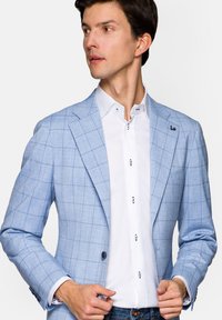 Jasnoniebieska marynarka w kratę wykonana z lekkiego materiału, zestawiona z białą koszulą z niebieskimi akcentami i detalami guzików. Widoczne dżinsy.