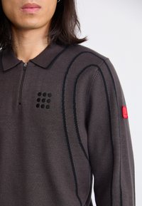 Temno siva polo majica z zip ovratnikom, ki ima črne šive in rdeč poudarek na rokavu. Mehka, teksturirana tkanina.