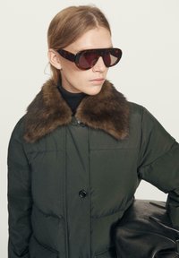 Woman with light brown hair wearing oversized tortoiseshell sunglasses and a dark puffer jacket with a brown fur collar, holding a black leather bag.