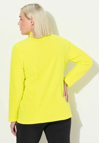 Keltainen fleece-takki, jossa on korkea kaulus, pitkät hihat ja sivusaumat, näyttää sileältä ja rentolta.