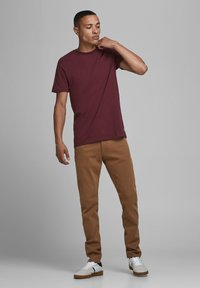 Bordeauxrotes Baumwoll-T-Shirt kombiniert mit hellbraunen Hosen. Das Model steht entspannt da und trägt weiße Sneakers mit navyfarbenen Akzenten. Einfaches Design, ohne Muster.