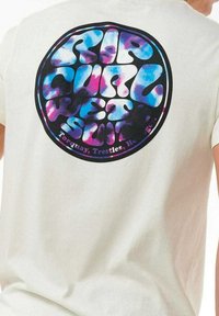 Biała bawełniana koszulka z okrągłą grafiką w kolorach niebieskim, różowym i czarnym oraz wyraźnym napisem: "RIP CURL WETSUIT" z szczegółami lokalizacji poniżej.