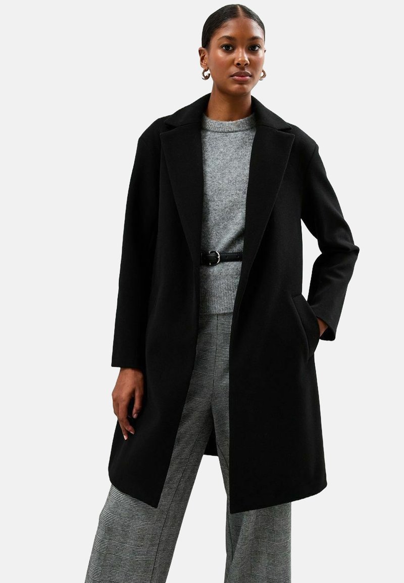 Zwarte wollen jas met een gekarteld colbert, gestructureerde silhouet en voorzakken. Ggedragen over een lichtgrijze trui en geblokte broek.