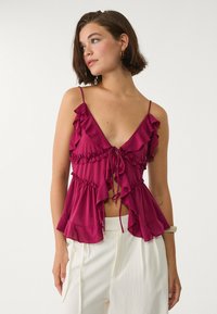 Fuchsia ärmlös topp med volangdetaljer, djup V-ringning och åtsittande midja, i kombination med vita byxor. Slät textur, lättviktig tyg.