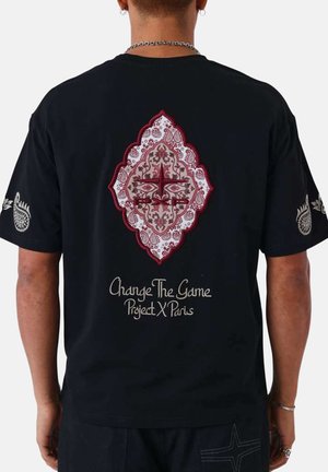 Homme portant un t-shirt noir avec un motif en forme de losange paisley rouge et blanc et le texte « Change The Game Project X Paris » au dos.