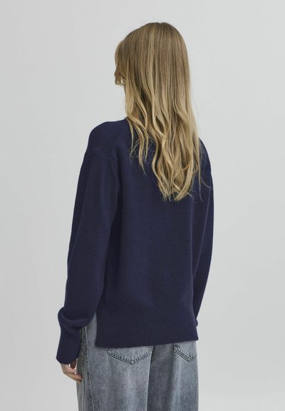 Mørk marineblå strikket genser med avslappet passform, ribbestrikkede mansjetter og kant, sammen med lysegrå løse jeans. Bakside, glatt tekstur.