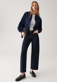 Marine bomberjack, witte aansluitende top, donkere denim wijde broek, zwarte loafers en een kleine zwarte crossbody tas. Gladde texturen en casual pasvorm.