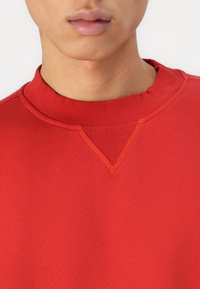 Rød hettegenser med ribbestrikket rund hals, som har en V-formet sømdetalj ved kragen. Materialet ser glatt og lett ut.