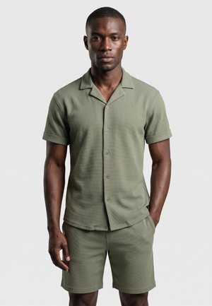 Homme portant une chemise boutonnée à manches courtes et un short assortis de couleur vert olive, debout sur un fond clair uni.
