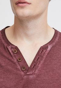 Burgundy-koska T-paita V-halkiolla, jossa on kolme puukuvioista nappia ja kontrastivärinen ompelu kauluksen reunassa. Yleisvaikutelma on sileä.