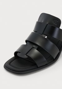 Sandale glissante en cuir noir avec une semelle plate, trois larges sangles disposées en design tissé, avec une texture lisse et un subtil détail de logo.