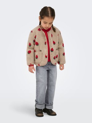 Jeune fille aux cheveux tressés portant une veste en polaire beige avec un motif floral rouge, un t-shirt rouge, un jean gris retroussé et des bottes noires, regardant vers le bas.