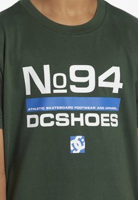Zelené tričko s krátkým rukávem s výrazným bílým a modrým textem: "No94" a "DCSHOES", s dalšími logy na látce.