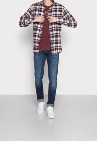 Man som knäpper en burgunderrandig skjorta över en burgundygrafisk t-shirt, iförd blå upprullade jeans och vita sneakers, stående inomhus.