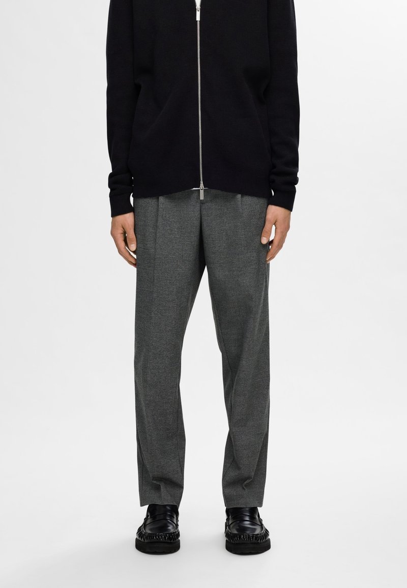 Svart stickad cardigan med dragkedja och långa ärmar, tillsammans med grå houndstooth-byxor och svarta läderskor med fransdetaljer.