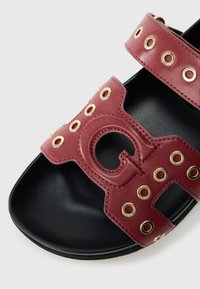 Sandale en cuir rouge avec un design à lanières, œillets dorés et un logo découpé bien visible. Semelle noire à texture lisse.