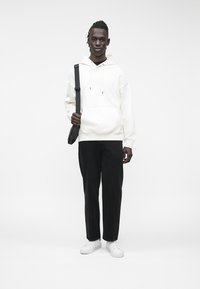 Sweat à capuche blanc avec cordons de serrage, pantalon noir, et baskets blanches. Le modèle tient un sac à bandoulière noir, debout devant un fond clair.