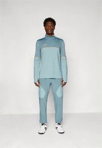 Férfi áll, világoskék sportos melegítő felsőt visel narancssárga szegéllyel és fehér sportcipőben, egyszínű háttér előtt.