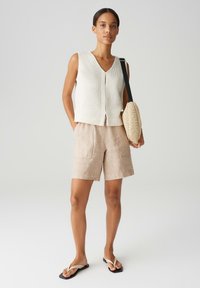 Top senza maniche in cotone avorio, pantaloni corti in lino beige chiaro con tasche e infradito nere. La modella tiene una borsa tessuta e strutturata.