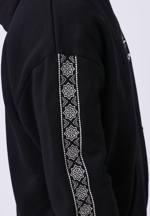 Vêtement noir à manches longues avec une bande à motif géométrique blanc courant verticalement le long de l'extérieur du bras.