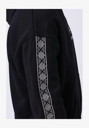 Vêtement noir à manches longues avec une bande à motif géométrique blanc courant verticalement le long de l'extérieur du bras.