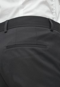 Calças cinza escuras com uma textura suave, apresentando um único bolso traseiro e passantes para cinto, combinadas com uma camisa branca na parte superior.