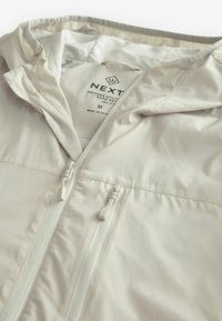 Lightweight cream jacket with a hood, features a full front zipper, elastic cuffs, and a smooth texture. Label tags visible inside.