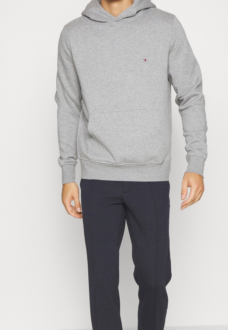 Felpa grigia in cotone con tasca a marsupio frontale, polsini e orlo a costine, con un piccolo logo sul petto. Indossata con pantaloni blu navy.
