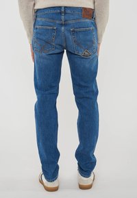 Uomo che indossa jeans di denim blu con tasche posteriori e una toppa in pelle marrone, abbinati a sneakers bianche e un maglione beige, in piedi di spalle.
