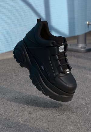 Czarny masywny sneaker z grubą podeszwą i logo „Buffalo”, umieszczony na teksturowanej powierzchni asfaltu w pobliżu jasnoniebieskiej ściany i metalowego balustrady.