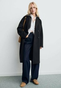 Abrigo negro de doble botonadura hecho de una mezcla de lana, usado sobre una camisa blanca, combinado con jeans de pierna ancha y mocasines marrones. Bolso color tostado.
