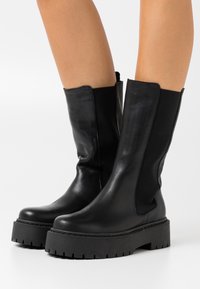Svarta läderankelboots med elastiska sidopaneler, kraftiga svarta gummisulor och en slät finish. Texturerade för grepp och stabilitet.