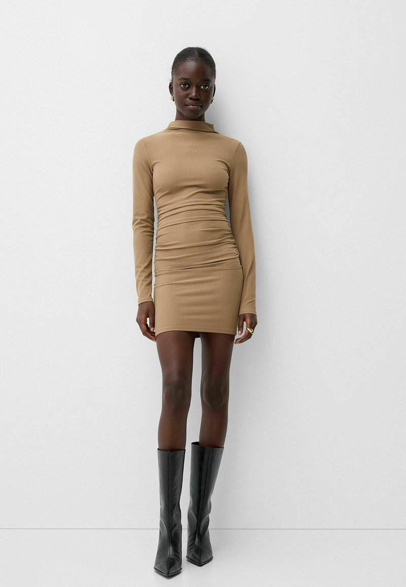 Donna che indossa un mini abito lungo attillato color beige con arricciature sui lati, abbinato a stivali neri a punta al ginocchio, in piedi contro un muro bianco.