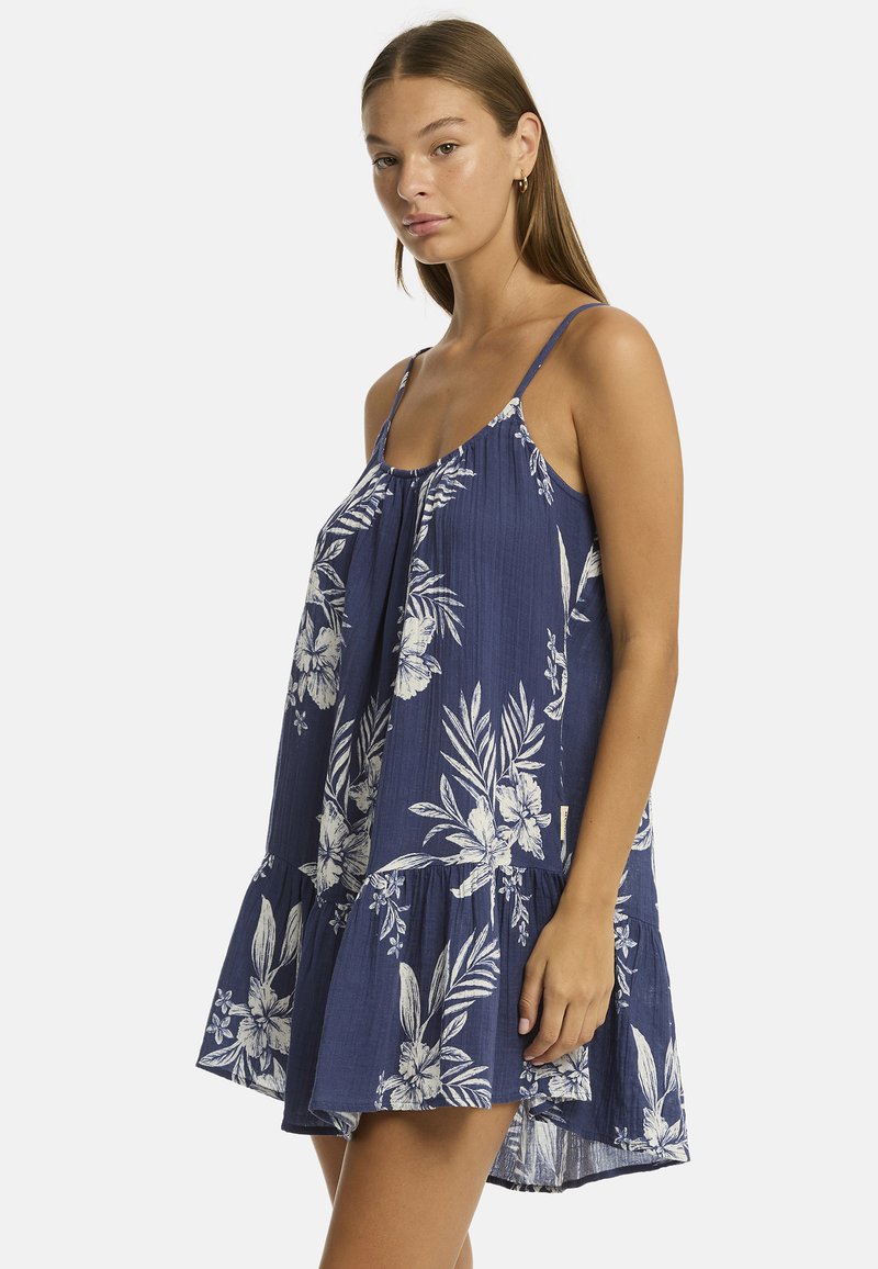 La modelo lleva un vestido floral azul con un dobladillo acampanado, que cuenta con tirantes ajustables y un corte holgado, posando frente a un fondo liso.