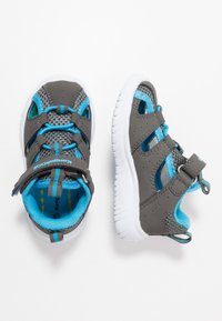 Grå joggesko med blå detaljer, mesh-overdel, hvit gummisåle, justerbar reim og flere ventilasjonshull for pusteevne.