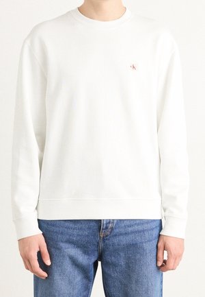 Osoba w prostym białym sweatshirtcie z okrągłym dekoltem i małym czerwonym logo "CK" na piersi, w połączeniu z niebieskimi jeansami.