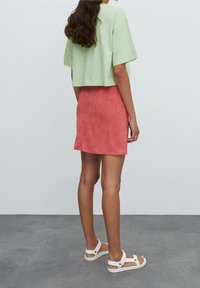 Camiseta de algodón verde claro, recortada, combinada con una mini falda de ante rosa. Modelo con sandalias planas blancas, mostrando las piernas y un estilo casual.