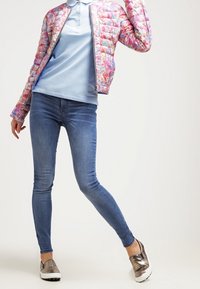 Vadderad jacka med blommönster i rosa och blått, ljusblå pikétröja, slim fit blå jeans och metalliska slip-on skor.