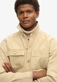 Chaqueta de lona beige con un cuello alto, dos bolsillos frontales y detalles en hardware dorado. Llevada sobre un suéter de canalé beige claro.