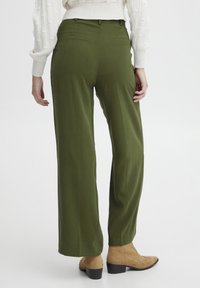 Pantaloni a gamba larga verde oliva con una texture liscia, due tasche posteriori e una vestibilità su misura, abbinati a stivali alla caviglia color beige chiaro.
