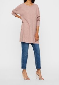 Frau in einem langen pinkfarbenen Pullover, blauen hochgekrempelten Jeans und silbernen High-Heels, die mit einer Hand an der Hüfte steht.