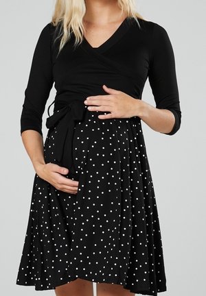 Gravid kvinde i en sort omslagskjole med hvide polkaprikker, der blidt holder sin mave med begge hænder.